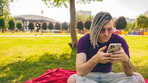 Um jovem com dreadlocks está sentado em um cobertor no parque e usando um smartphone Um cara bonito está sentado em um gramado verde e assistindo conteúdo no telefone em um dia ensolarado