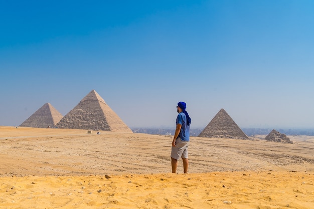 Um jovem com um turbante azul caminhando ao lado das Pirâmides de Gizé, o monumento funerário mais antigo do mundo. Na cidade do Cairo, Egito