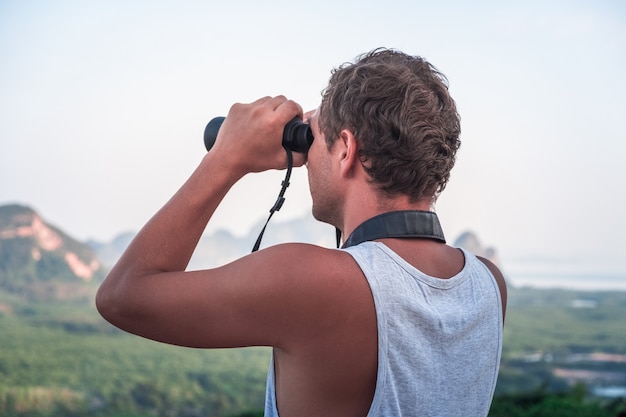 Um jovem com uma camiseta branca olha de cima para baixo com binóculos para a vida selvagem.