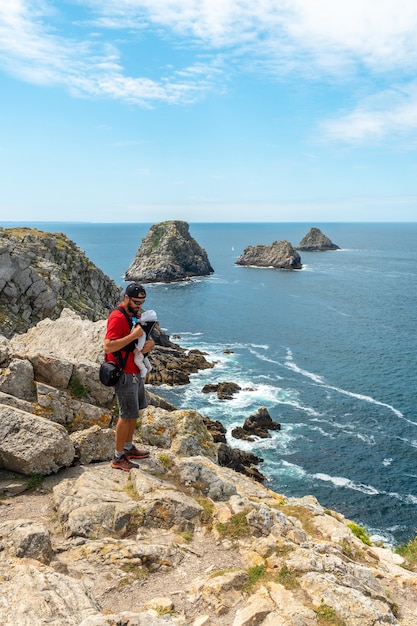 Um jovem com uma mochila e um bebê olhando o mar em pen hir point na península de crozon, na bretanha francesa, as três ilhotas famosas, frança