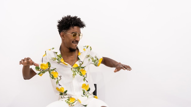 Um jovem cubano com cabelo encaracolado, sentado em uma camisa branca de flores e óculos. Sorrindo e dançando em um fundo branco