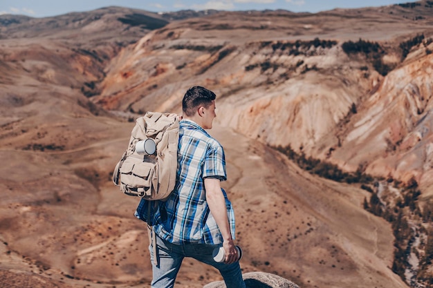 Um jovem de camisa xadrez com uma mochila de caminhada e uma garrafa térmica na mão descansa o pé em um paralelepípedo apreciando a vista do cânion. O conceito de turismo.