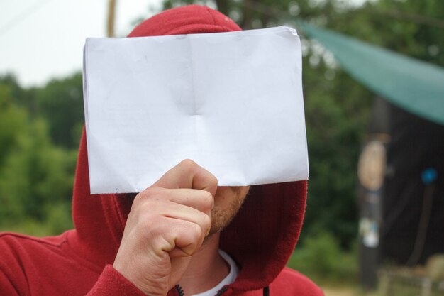 Um jovem de capuz vermelho detém uma folha de papel branco na frente de seu rosto.