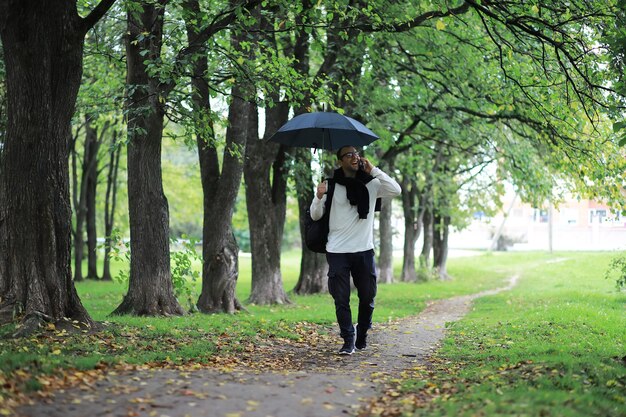 Um jovem de óculos anda no parque com um guarda-chuva durante a chuva.