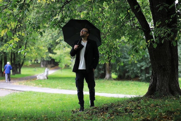 Um jovem de óculos anda no parque com um guarda-chuva durante a chuva.