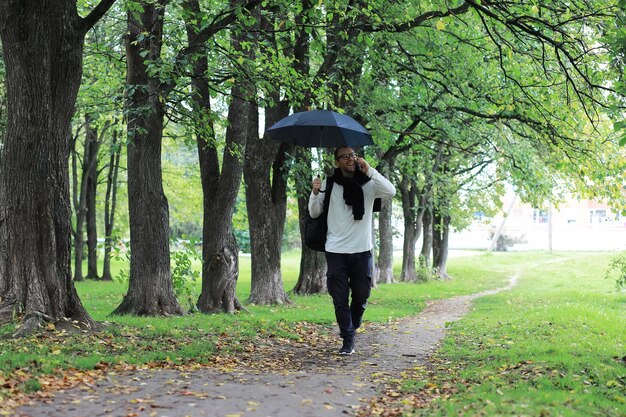 Um jovem de óculos caminha no parque com um guarda-chuva durante a chuva