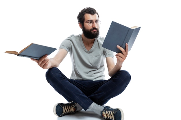 Um jovem de óculos e barba senta-se em uma pose de ioga e tem livros nas mãos. Educação e treinamento. Isolado.