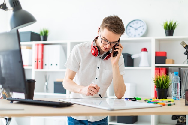 Um jovem de óculos fica perto de uma mesa de computador e está falando ao telefone. diante dele está um quadro magnético e marcadores. no pescoço, os fones de ouvido do cara estão pendurados.