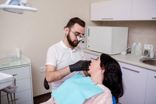 Um jovem dentista especialista examina e trata os dentes de uma bela mulher na odontologia branca moderna usando um microscópio Tratamento de próteses dentárias e clareamento dos dentes