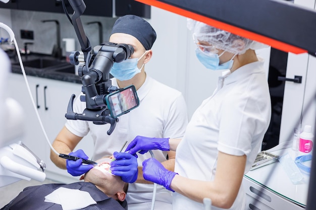 Um jovem dentista olha para os dentes do paciente com um microscópio odontológico e segura instrumentos dentários perto da boca Um assistente ajuda o médico Eles usam uniformes brancos com máscaras