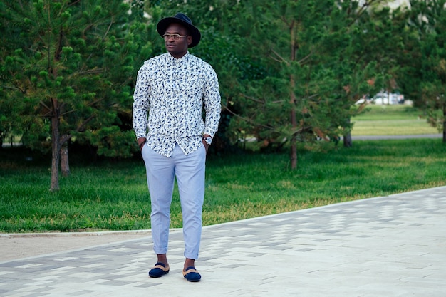 Um jovem e bonito modelo elegante homem afro-americano em um elegante terno e um chapéu preto em um parque de verão. empresário latino-americano hispânico negro posando para a sessão de fotos