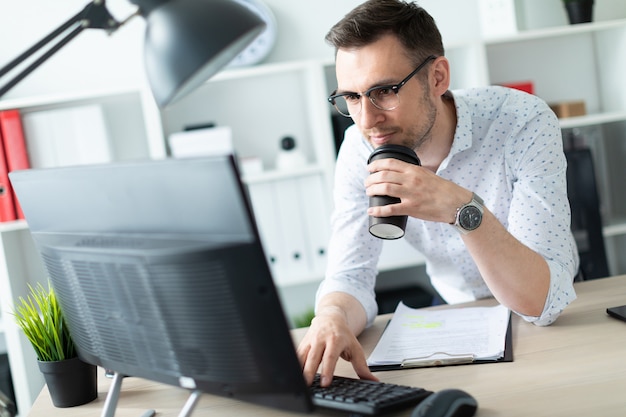 Um jovem em copos fica perto de uma mesa no escritório, detém um copo de café na mão e trabalha com um computador