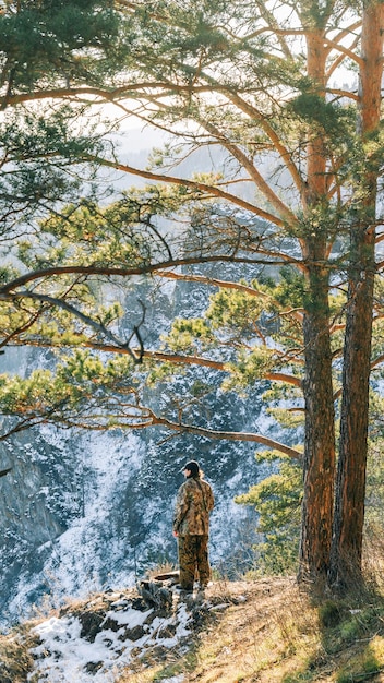 Um jovem em roupas de camuflagem fica em uma encosta ensolarada de uma rocha contra o pano de fundo das montanhas cobertas de neve