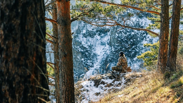 Um jovem em roupas de camuflagem senta-se em um tronco em uma encosta ensolarada de uma rocha contra o pano de fundo das montanhas cobertas de neve