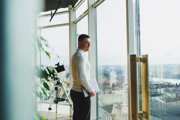 Um jovem em um espaço de trabalho moderno fica perto de uma grande janela Um homem em roupas casuais com um laptop nas mãos Trabalho remoto Bom jovem gerente