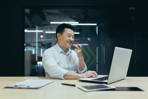 Um jovem empresário asiático que trabalha no escritório com laptop e documentos fala ao telefone através