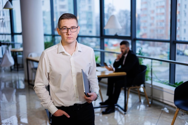 Um jovem empresário de óculos e camisa branca com um laptop nas mãos fica no escritório e trabalha