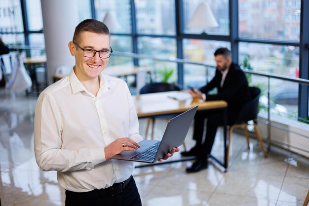 Um jovem empresário de óculos e uma camisa branca com um laptop nas mãos fica no escritório e trabalha