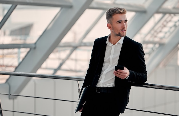 Um jovem empresário em um centro de negócios com um telefone e um laptop na mão em um terno preto e camisa branca olhando para longe