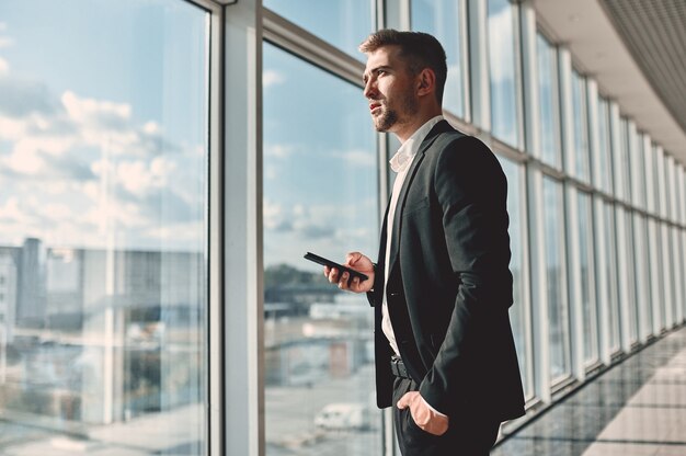 Um jovem empresário em um centro de negócios em um terno preto e camisa branca com um telefone na mão e a outra no bolso, olhando pela janela