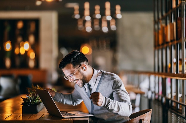 Um jovem empresário feliz está comemorando a vitória enquanto ri do laptop na cafeteria