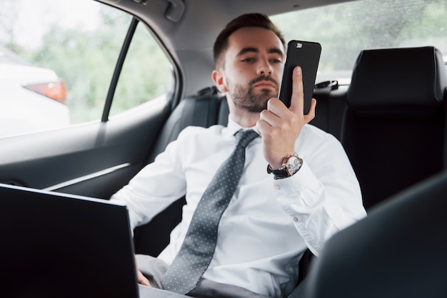 Um jovem empresário trabalhando no laptop e falando ao telefone enquanto está sentado na parte de trás do carro. Trabalha em movimento, aprecia seu tempo