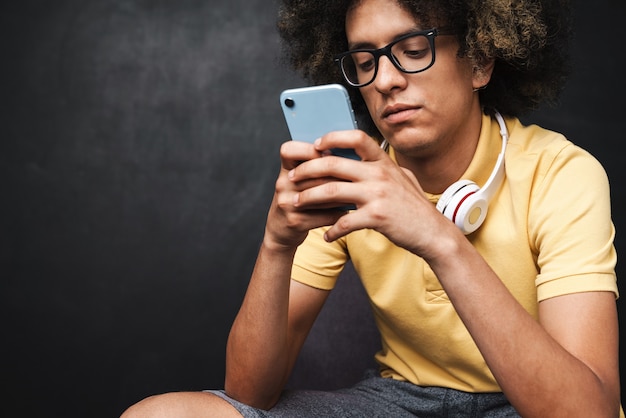 um jovem encaracolado sério adolescente concentrado posando sobre uma parede de lousa cinza com fones de ouvido usando telefone celular.