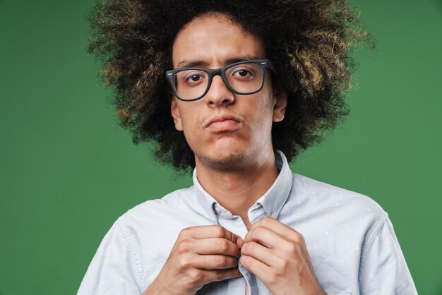 Um jovem encaracolado sério posando isolado na parede verde, vestido com uma camisa branca formal.