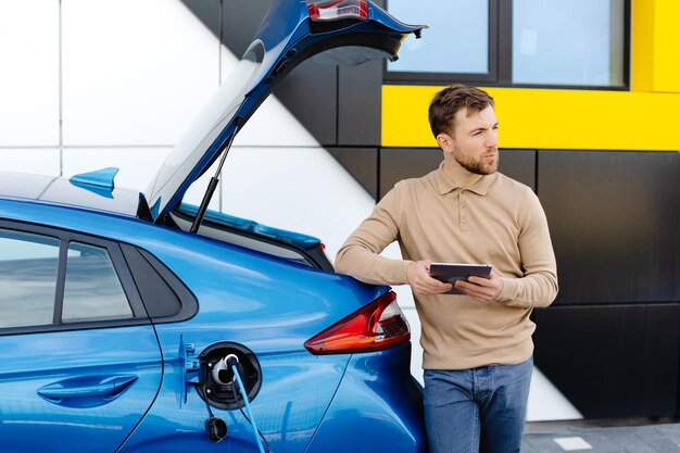 Um jovem está carregando um carro elétrico enquanto está parado em uma estação e segurando um tablet