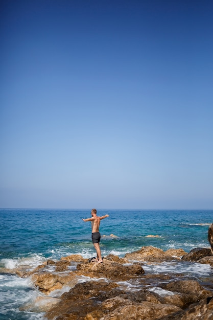 Um jovem está de pé nas rochas com vista para o mar Mediterrâneo aberto. Um cara em um dia quente de sol de verão olha para a brisa do mar