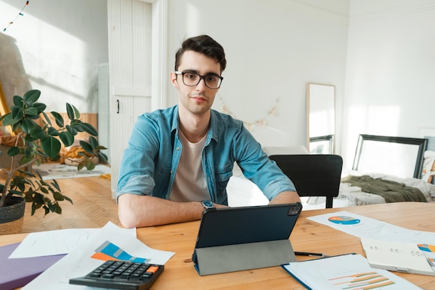 Um jovem está digitando em um laptop contando com uma calculadora e trabalhando com documentos em casa Olhando para a câmera