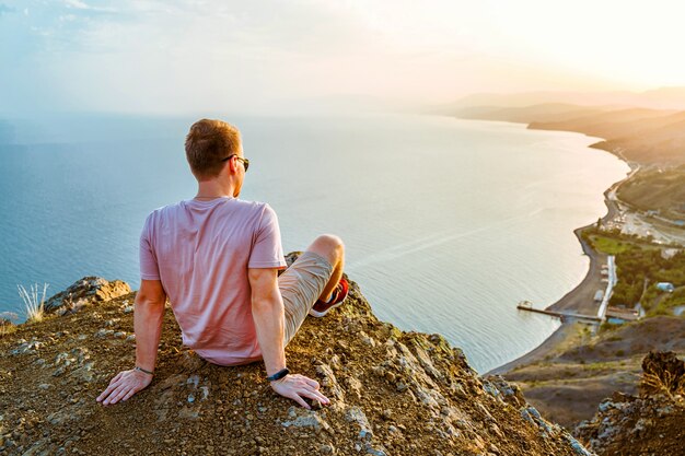 Um jovem está na beira de um penhasco ao pôr do sol com uma vista incrível das montanhas