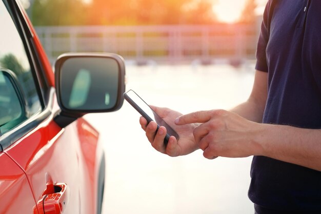 Um jovem está parado perto de seu carro usando um smartphone ao pôr do sol