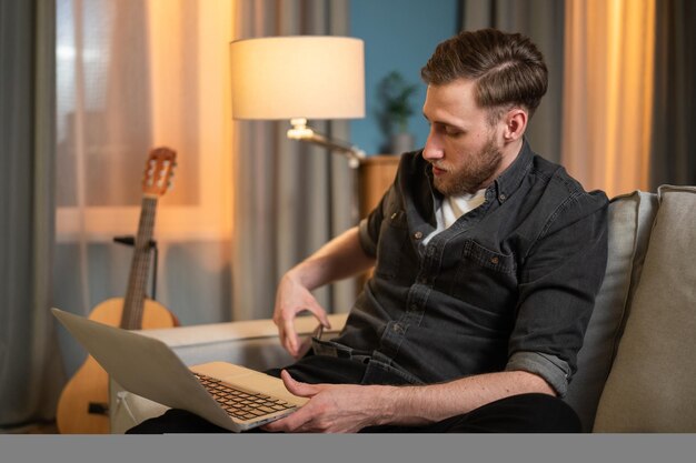 Um jovem está relaxando em um sofá com um laptop no colo, a mão pega o telefone que ele tira