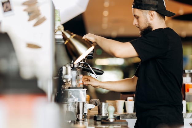 Um jovem estiloso, com barba, vestindo roupas casuais, cozinha café em uma cafeteira em uma cafeteria aconchegante. .