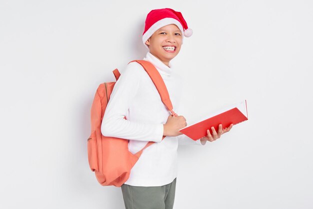 Um jovem estudante asiático alegre e bonito com um chapéu de Natal vestindo uma camiseta com mochila segurando livro isolado em fundo branco