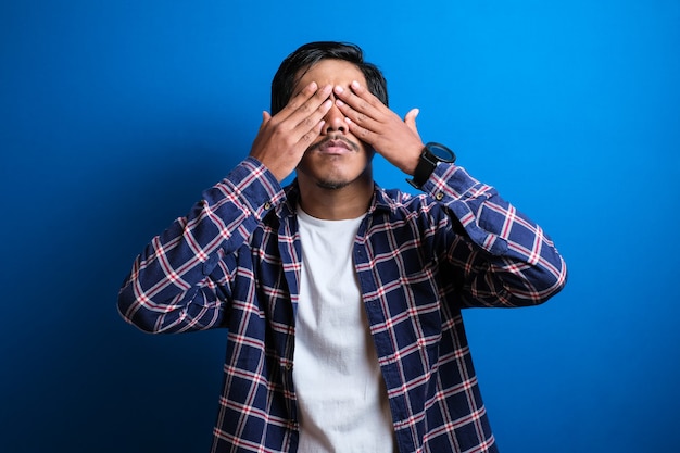 Um jovem estudante fechou os olhos com as duas mãos contra um fundo azul