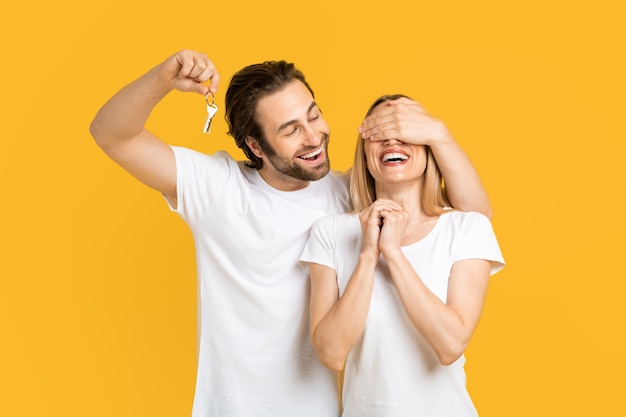 Foto um jovem europeu sorridente em camiseta branca fecha os olhos para sua esposa e segura chaves isoladas em amarelo