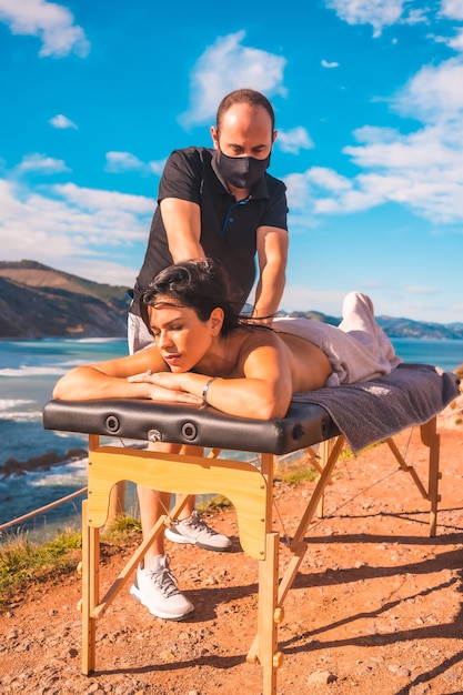 Um jovem fazendo uma massagem na natureza na costa perto do mar, massagista com máscara facial na pandemia de coronavírus