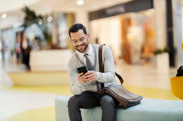 Um jovem feliz em casual inteligente está sentado em um banco no shopping e mensagens de texto