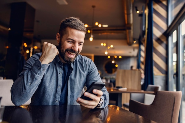 Um jovem feliz está sentado comemorando a vitória na cafeteria enquanto olha para o telefone