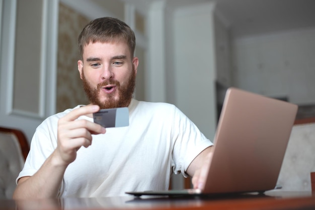 Um jovem feliz segura o cartão do banco de crédito na mão, usa o sorriso do computador portátil em casa, fazendo compras on-line