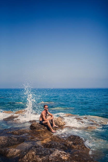 Um jovem fica nas rochas com vista para o mar Mediterrâneo aberto Um cara em um dia quente e ensolarado de verão olha para a brisa do mar