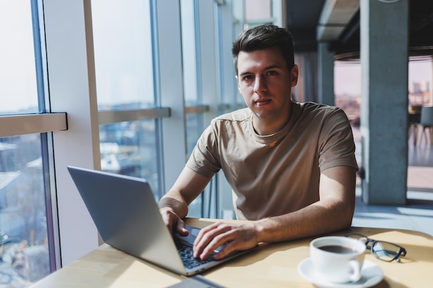 Um jovem freelancer masculino atraente senta-se no interior de uma cafeteria e olha pela janela Blogueiro bonito escrevendo ideias no laptop enquanto está sentado na mesa de madeira no café com laptop