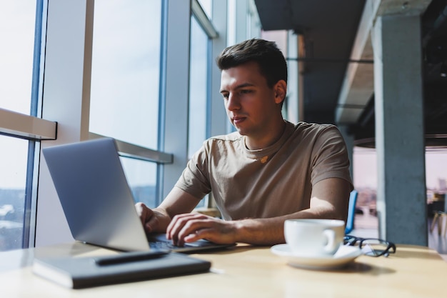 Um jovem freelancer masculino atraente senta-se no interior de uma cafeteria e olha pela janela Blogueiro bonito escrevendo ideias no laptop enquanto está sentado na mesa de madeira no café com laptop