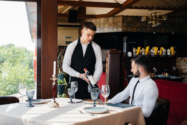 Um jovem garçom com um avental estiloso demonstra e oferece um bom vinho a um cliente em um restaurante. Atendimento ao Cliente.