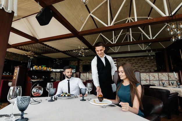 Um jovem garçom em um avental elegante serve uma mesa com um lindo casal em um restaurante refinado Delicadezas requintadas da alta cozinha closeup