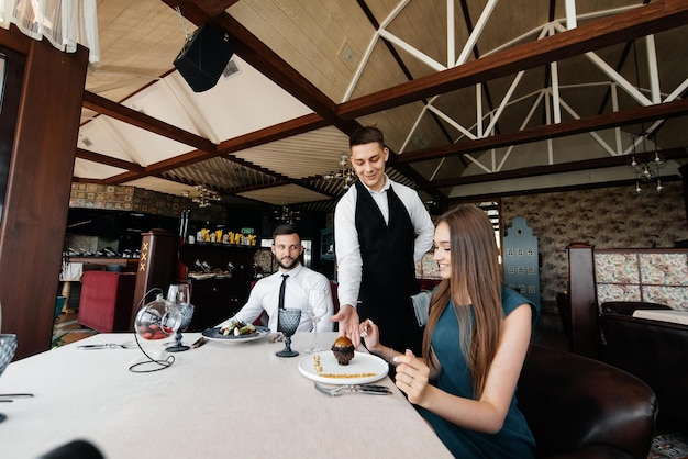 Um jovem garçom em um avental elegante serve uma mesa com um lindo casal em um restaurante refinado Delicadezas requintadas da alta cozinha closeup