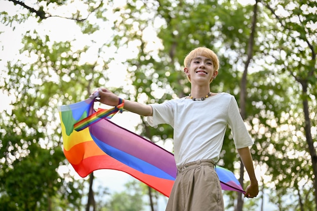 Um jovem gay asiático despreocupado com uma bandeira de arco-íris lgbt parada no parque verde