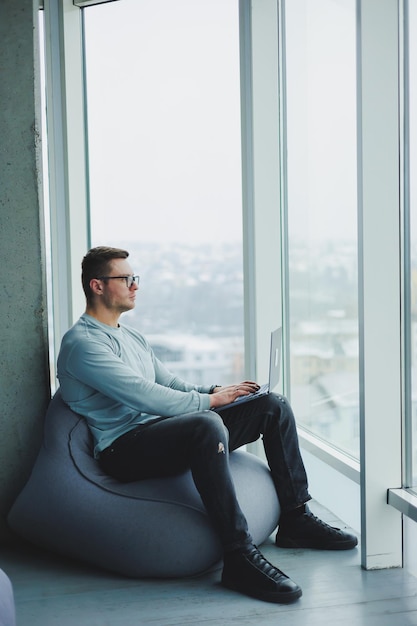 Um jovem gerente de óculos e roupas casuais senta-se em uma cadeira macia e trabalha em um laptop Espaço de trabalho para trabalho remoto